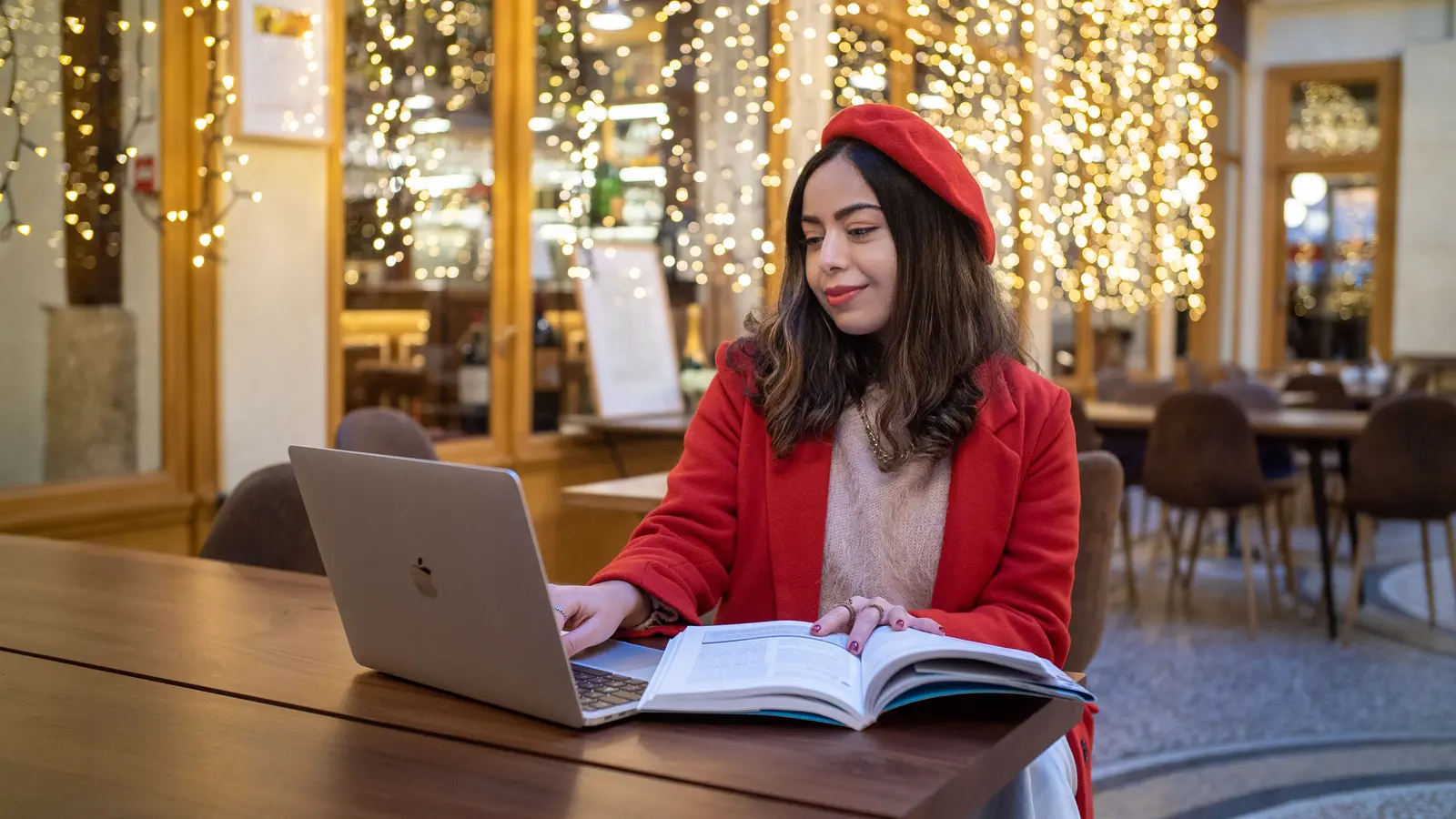 The best places to study in Paris Young woman in red beret studying with laptop in a Parisian café with festive lights.